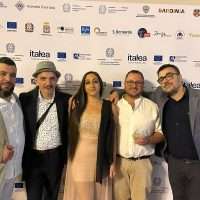 Gruppo di cinque persone sorridenti, evento formale, sfondo con logo sponsor e branding, atmosfera professionale e calorosa.