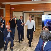 Uomo in camicia bianca entra in una sala conferenze, applaude mentre altri partecipano a un evento formale in ambiente elegante.