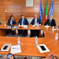 Riunione formale tra cinque professionisti seduti attorno a un tavolo in legno, con bandiere e documenti, aria di discussione.