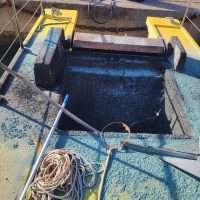Immagine di un pozzo o serbatoio su una piattaforma gialla, con riduzione metallica, cavi lasciati sul pavimento e riflessi d’acqua scura all’interno.