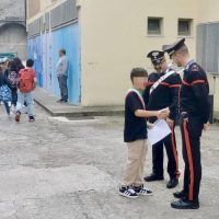 Bambino che riceve un certificato da due militari in uniforme, in cortile urbano con costruzioni colorate sullo sfondo.