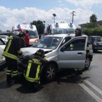 Soccorritori e vigili del fuoco pronti all’intervento accanto a un’auto incidentata in strada, con sirene in background.