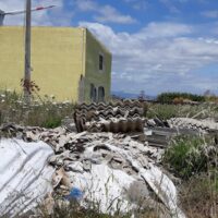 Cantina di cemento gialla sullo sfondo, rocciose pile di pneumatici e rifiuti rotti in primo piano, cielo azzurro.