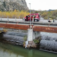 Auto rossa parcheggiata su strada, ponte in primo piano con corrugamenti d’acqua e persona sotto a un ponte; paesaggio collinare e alberi autunnali sullo sfondo.