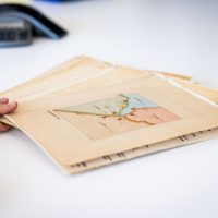 La seconda mano tiene foglianti antichi con disegno tecnico su sfondo bianco, atmosfera di lavoro creativo e archivio cartaceo.