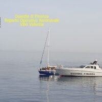 Barca a vela al largo con motoscafo, mare calmo e cielo luminoso; testo giallo in alto sinistra: 'Cooperativa ...', scena marina tranquilla.