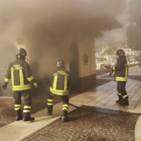 Squadre dei vigili del fuoco in azione davanti a un edificio avvolto da fumo, indossano tute nere e protezioni gialle, intervento in corso.