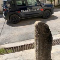 Auto dei carabinieri sullo sfondo, marciapiede e una vecchia colonna di pietra al primo piano.