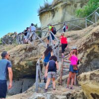 Gruppo di persone che saluta e cammina su una passerella di legno su una scogliera, sfondo mare e cielo azzurro.