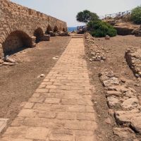 Sentiero lastricato tra resti di mura e archi antichi, con il mare sullo sfondo e cespugli verdi ai lati.