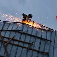 Operaio al lavoro su una tettoia in metallo; scintille e fuoco generano bagliori arancioni contro il cielo, visibile a lato.