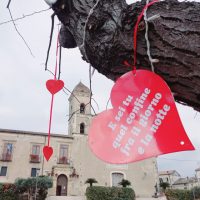 Cuori rossi pendenti da un albero in primo piano, con chiesa di pietra sullo sfondo, evocando amore e romanticismo.