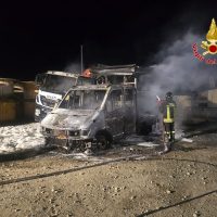 Autoveicolo ucrivo gravemente danneggiato da incendio notturno, vigili del fuoco presenti vicino al veicolo fumante, scena di recupero.