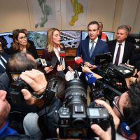 Un gruppo di politici in conferenza stampa circondati da giornalisti e giornalisti, con microfoni e telecamere, atmosfera di immediato reportage.