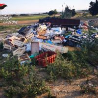 Rifiuti abbandonati e pattume sparso in un campo, con cassette e materiali vari tra cespugli e terra, scena di degrado ambientale.