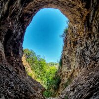 Vista dall'interno di una grotta verso il cielo azzurro, circondata da pareti rocciose e vegetazione luminosa all'aperto.