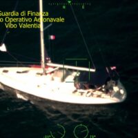 Luna di notte: barca a vela sul mare, luci verdi sull’oceano, testo sovrapposto in alto a sinistra. Scena marina investigativa.