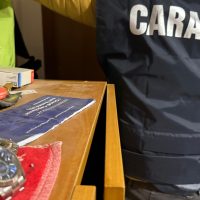 Scena di una tastiera e di una manica di giubotto con scritte, angolo di tavolo con oggetti vari; presente figura in background.