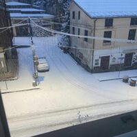 Vista notturna o serale di una strada innevata tra case e potature: tetto e viario coperti da neve, luci accese.