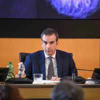 Uomo in abito parla al microfono in una conferenza, tavolo con bottiglia d’acqua e auditorio sullo sfondo.