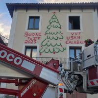 Edificio con grande albero di Natale dipinto all’esterno, finestre aperte e usci segnati, vigili del fuoco con mezzo presente davanti all’ingresso.