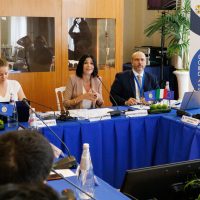 Riunione formale in sala conferenze: tre relatori seduti al tavolo con badge, documenti e laptop, pubblico sullo sfondo.