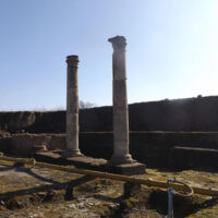 Due colonne antiche in rovina tra resti di mosaici e mura di pietra, cielo azzurro e luce solare che illumina il sito archeologico.