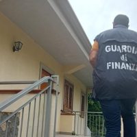 Guardia di Finanza in abito civile controlla l’ingresso di un edificio residenziale, inquadratura laterale dall’esterno.