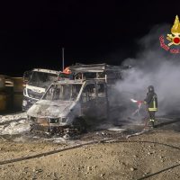Autobus incendiato sulla strada al buio, fumo visibile e vigili del fuoco che gestiscono l’emergenza.