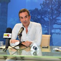 Uomo in abito chiaro parla al microfono durante una trasmissione radiofonica in studio, con borracce e tazze sul tavolo.