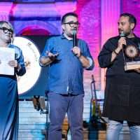 Tre uomini sul palco durante una presentazione: due con microfono parlano, uno con cartello e occhiali, sfondo colorato e strumentazione musicale.