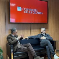 Due uomini seduti su divani neri discutono in una sala conferenze; sullo sfondo uno schermo rosso con logo Università della Calabria.