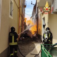 Vigili del fuoco in uniforme antisfiogo fronte a un incendio in una viuzza stretta, fiamme e fumo brillano tra edifici gialli.