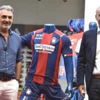 Allenatore o giocatore vicino a una maglia da calcio rosso e blu, con persone in background in un evento sportivo.