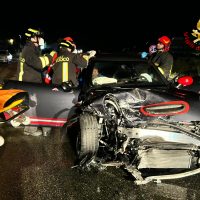 Grave incidente stradale notturno: auto frontalmente danneggiata, soccorritori in casco e giubbotti riflettenti soccorrono i feriti sull’asfalto