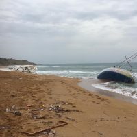 Spiaggia sabbiosa con riflessi d’acqua, motoscafo arenato e residui di onde; cielo nuvoloso e costa in lontananza.