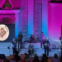 Concerto live su gradinate: musicisti su palco con luci rosa e blu, pubblico in prima fila, atmosfera festosa.