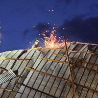 Lavoratori su una tettoia in legno mentre esplodono scintille e scintillano fuochi d'artificio, sfondo cielo serale blu.