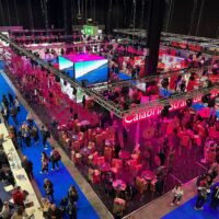 Grande evento fieristico internazionale con stand colorati e aree conferenze; visitatori in abiti vivaci percorrono corridoi rossi e blu, atmosfera dinamica.