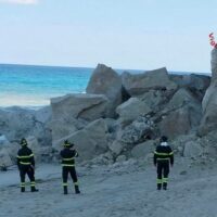 Tre vigili del fuoco in tuta gialla/nera fronteggiano una scogliera rocciosa su una spiaggia sabbiosa, mare azzurro sullo sfondo.