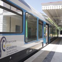 Treno regionale blu e bianco in stazione, vetrate e piattaforma soleggiata, logo Ferrovie Calabria sul lato.