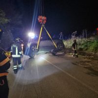 Intervento notturno su strada: uomini in divisa con cestello, gru solleva veicolo incidentato tra luci lampeggianti e fumo.