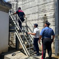 Tre poliziotti in uniforme sorvegliano un uomo mentre sale una scala metallica esterna in un cortile di blocco, con pareti di cemento.