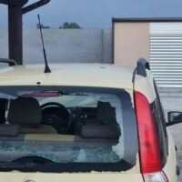 Auto beige con finestra posteriore infranta e vetro rotto su lato destro, parcheggiata in cortile con edificio sullo sfondo.