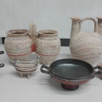 Vasi di ceramica grezza su tavolo, tonalità beige con motivi rossi; brocca e ciotola metallizzata presenti in primo piano.