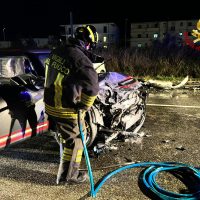 Incidente stradale notturno: vigili del fuoco davanti a veicolo danneggiato, attrezzature e cavi arrotolati a terra, ambiente urbano.