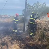Due vigili del fuoco in divisa riflessiva combattono un incendio su terreno sassoso, con fumo visibile e una parete di pietra sullo sfondo.