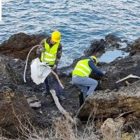 Due operai in giacca alta visibilità gialle lavorano su rocce costiere, con attrezzature e fascine lungo la costa rocciosa.