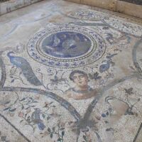 Mosaico antico con figure umane e motivi floreali, tonalità sabbia e blu, decorazioni circolari centrali.