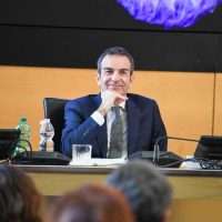 Uomo in abito seduto a una conferenza, sorridente, con microfono e bottiglia d'acqua sul tavolo.